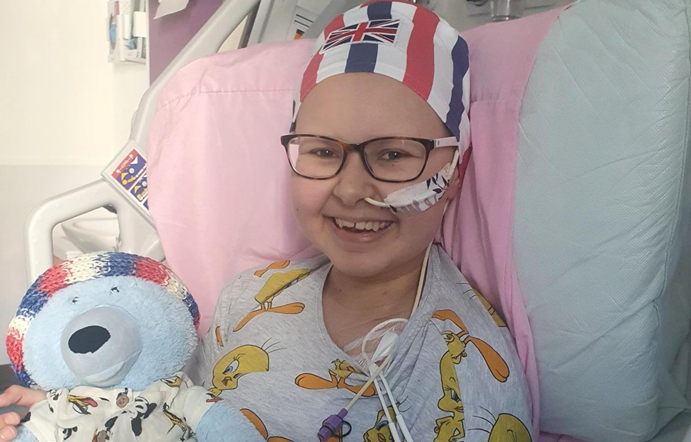 Teenage girl in hospital bed, holding teddy bear. She has a tube in her nose and wears a beanie.