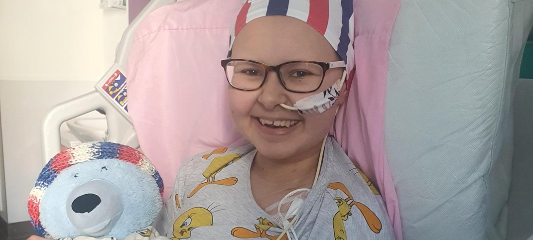 Teenage girl in hospital bed, holding teddy bear. She has a tube in her nose and wears a beanie.