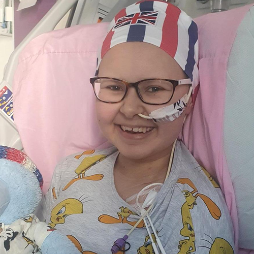 Teenage girl in hospital bed, holding teddy bear. She has a tube in her nose and wears a beanie.