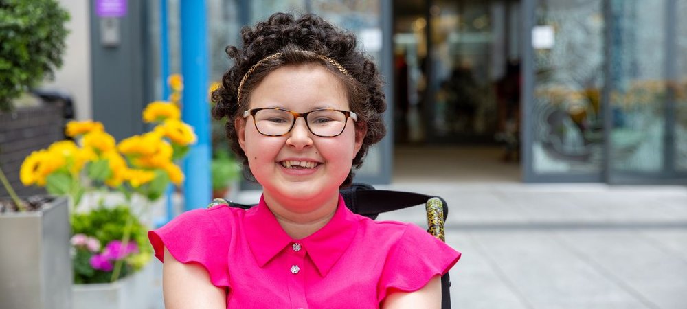 Teenage girl sitting in her wheelchair in front of GOSH entrance smiling