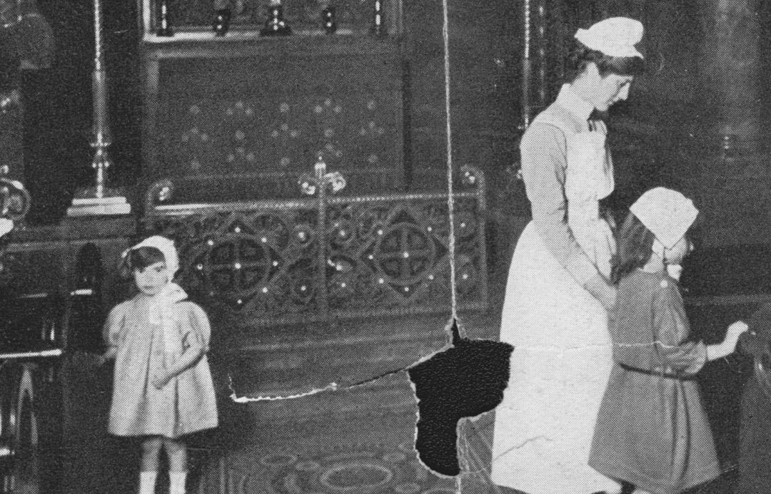 Black and white image showing two girls wearing bonnets inside the chapel. A nurse in floor length uniform stands with them. The image has been torn due to being old.