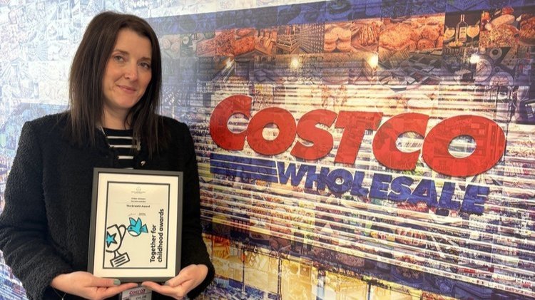 A woman holds an award certificate while standing next to a Costco sign.