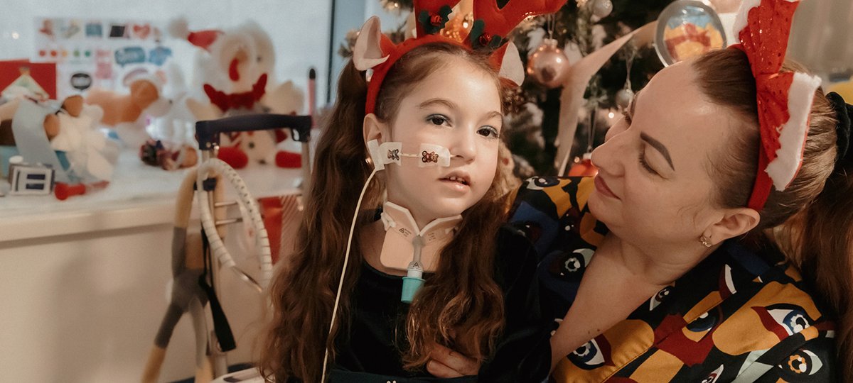 Girl sitting in her hospital room, next to her mum. They both wear festive hairbands.