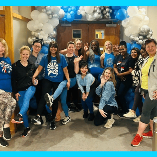 A group of 14 people posing and smiling in front of a balloon arch