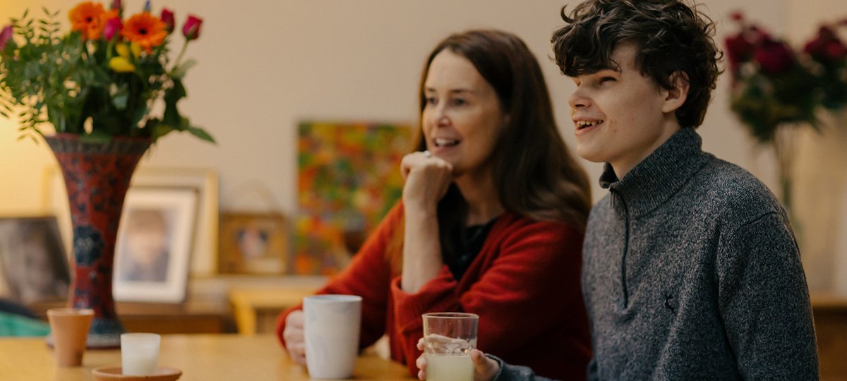 Teenage boy and his mum sit at kitchen table, holding drinks.
