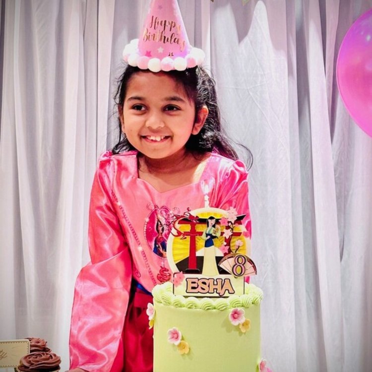 Little girl smiling. She is wearing a birthday hat and stands in front of a birthday cake with her name on it: Esha.