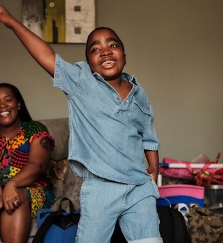 Little boy in his living room dancing. His mum sits behind him smiling.