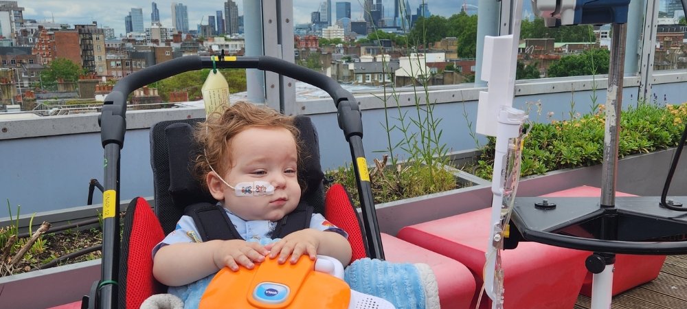 Little boy with feeding tube in buggy on rooftop garden. London skyline visible in the background.