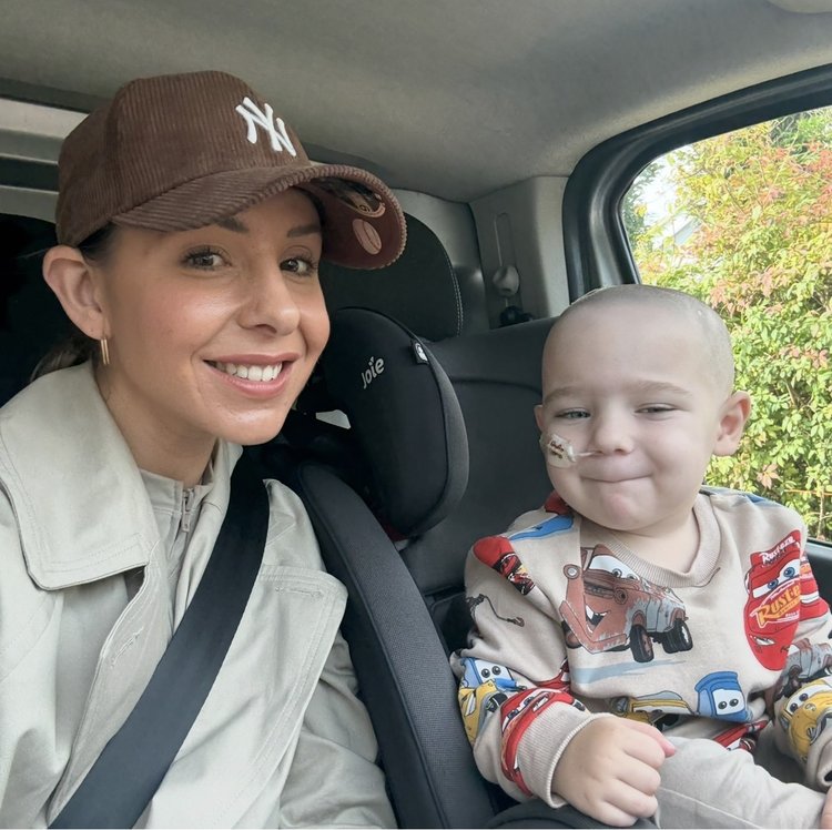Little boy sitting in car seat, smiling to camera. His mum sits next to him, smiling.