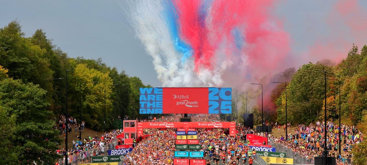 Image shows the beginning of the Great North Run, with thousands of runners crossing the start line. Behind them is the white, blue and red smoke from the Red Arrows air display