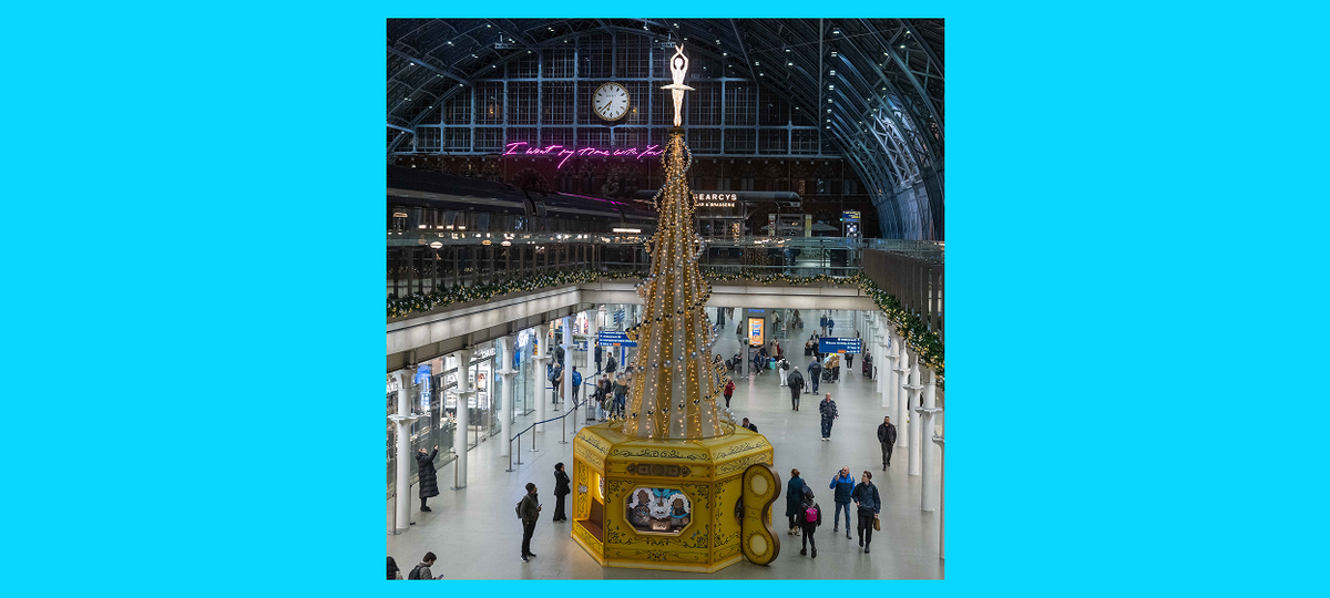 St Pancras X GOSH Charity Christmas tree 2025. It has an ornamental clockwork winder at the bottom, then baubles and lights decorate the tree itself, which is gold and white and shaped like a cone. An illuminated ballerina sits on the top.