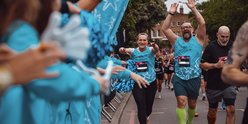 Two runners wearing blue GOSH running vests have their hands up in the air. They are running past a cheer point with people on the sidelines cheering them on