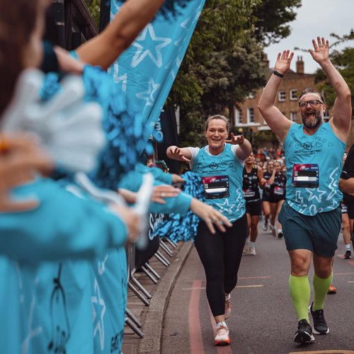 Two runners wearing blue GOSH running vests have their hands up in the air. They are running past a cheer point with people on the sidelines cheering them on