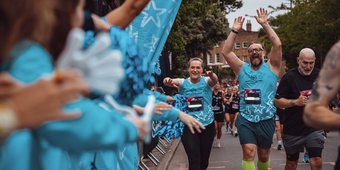 Two runners wearing blue GOSH running vests have their hands up in the air. They are running past a cheer point with people on the sidelines cheering them on