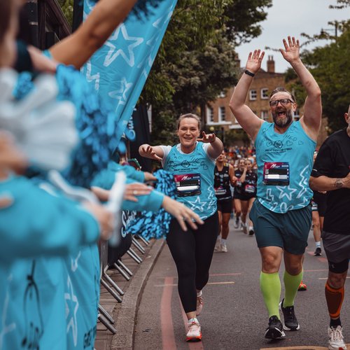 Two runners wearing blue GOSH running vests have their hands up in the air. They are running past a cheer point with people on the sidelines cheering them on