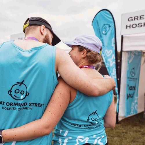 Two Hackney Half runners have their arms around each other, wearing blue GOSH branded running vests. The photo is taken from behind so we can't see their faces.