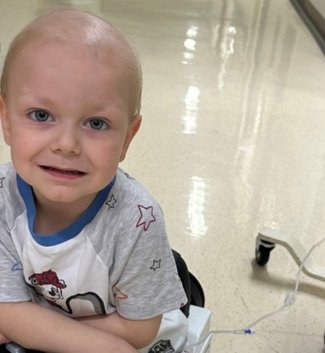 Close up of Hugo smiling and looking to camera. He's sitting in a toy car on an oncology ward, attached to a machine placed next to him..