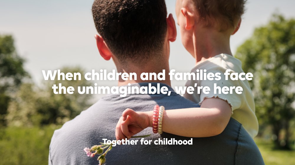 A father holds his child while standing in the park. The child is holding flowers. Text reads: When children and families face the unimaginable, we're here. Together for childhood.