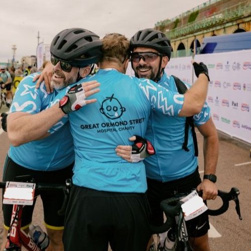 Three cyclists in GOSH branded cycling jerseys hug at the end of the London to Brighton cycle ride