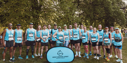 An image of a group of TeamGOSH runners before the start of the 2025 London Marathon. They are all wearing blue GOSH charity running vests. In front of them is a small banner. This is blue and has the GOSH Charity logo on it. The runners are stood infront of trees and are smiling to the camera