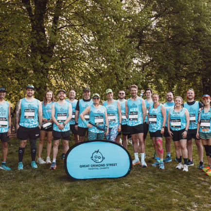 An image of a group of TeamGOSH runners before the start of the 2025 London Marathon. They are all wearing blue GOSH charity running vests. In front of them is a small banner. This is blue and has the GOSH Charity logo on it. The runners are stood infront of trees and are smiling to the camera
