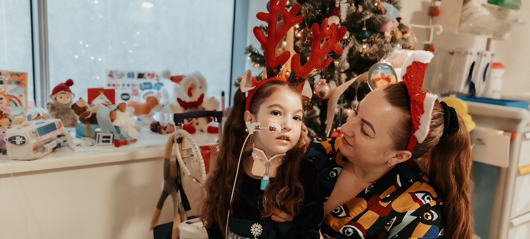 A girl wearing a hairband with reindeer antlers sits next to her mum. A Christmas tree stands behind them.