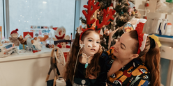 A girl wearing a hairband with reindeer antlers sits next to her mum. A Christmas tree stands behind them.