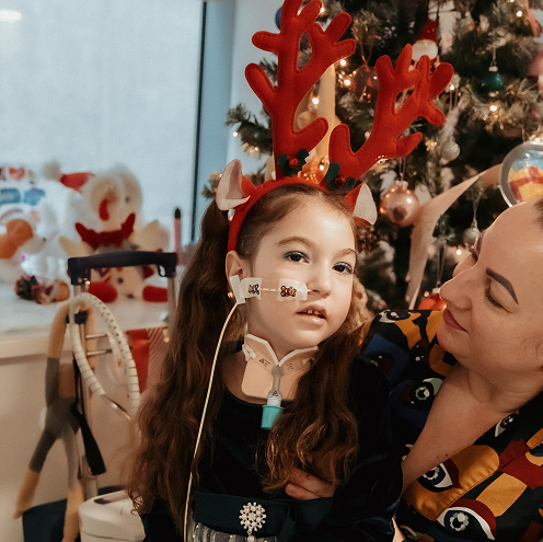 A girl wearing a hairband with reindeer antlers sits next to her mum. A Christmas tree stands behind them.