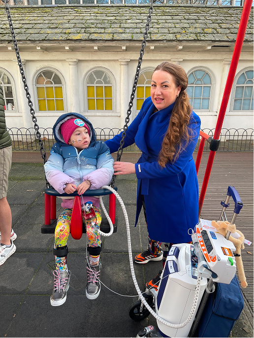 Maya on the swing in the park with her mum stood next to her.