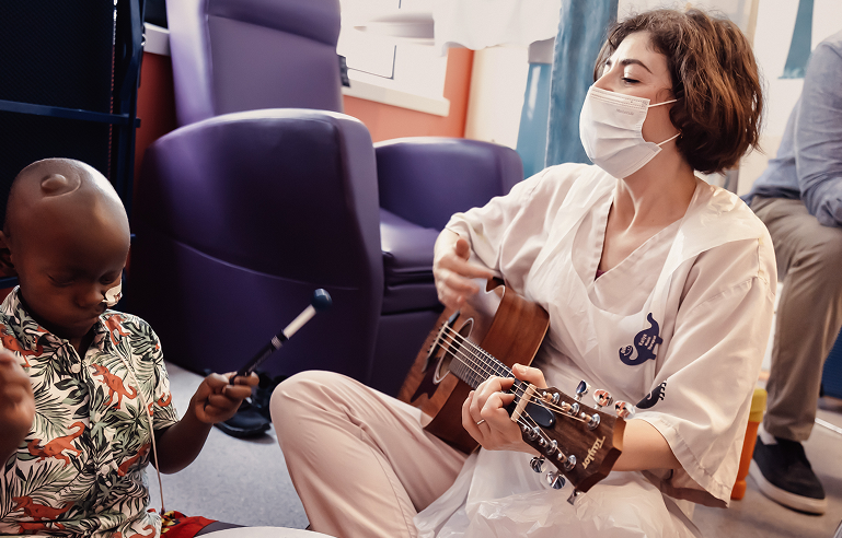 Music therapist Katya sitting playing a guitar. She is wearing a face mask. A little boy sits next to her, playing the drums.