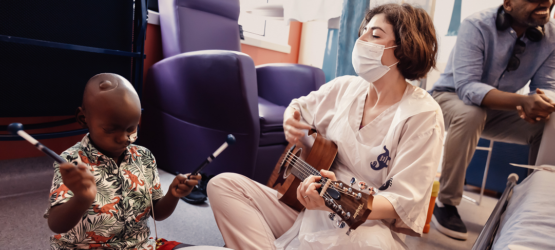 Music therapist Katya sitting playing a guitar. She is wearing a face mask. A little boy sits next to her, playing the drums.