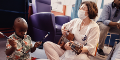 Music therapist Katya sitting playing a guitar. She is wearing a face mask. A little boy sits next to her, playing the drums.