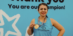 TeamGOSH runner standing in front of a blue background with the words 'Thank you. You are our champion' visible. The runner is smiling at the camera and showing the medal around their neck.