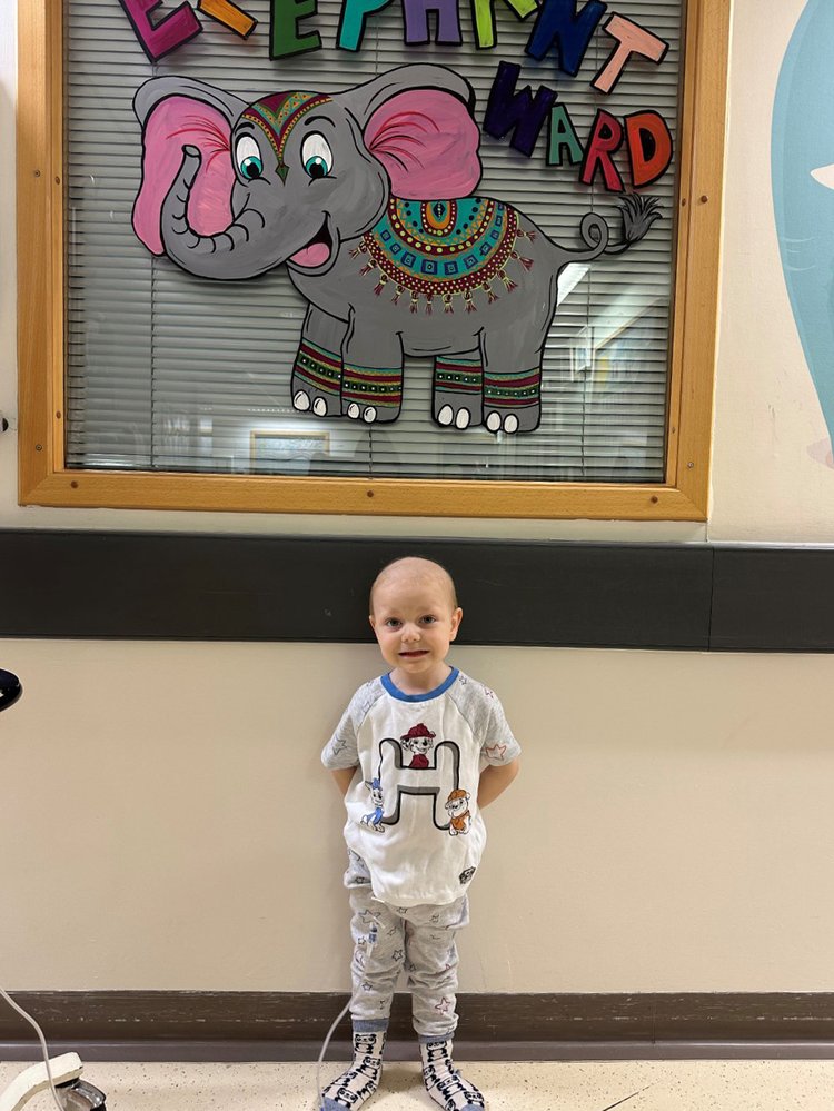 Hugo standing in front of a sign saying Elephant Ward. He's smiling and looking to camera. He is attached to a machine.