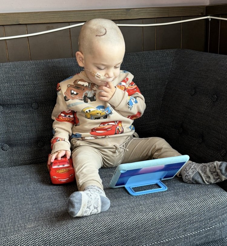 A little boy watching a tablet. He's wearing a Cars jumper and playing with a toy car