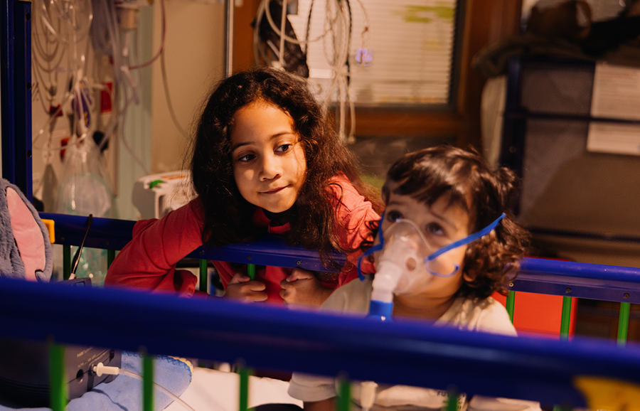 A little girl looking at her brother who is sitting in a hospital bed