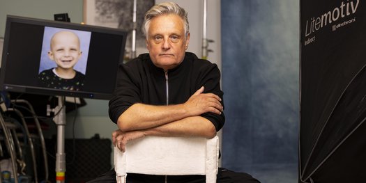 Photographer Rankin sits on a chair with his photos on a screen in the background