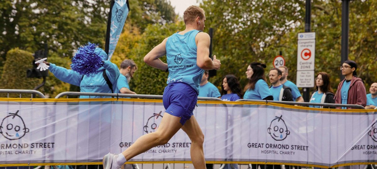 A runner wearing a blue GOSH Charity branded running vest runs past a white GOSH Charity banner
