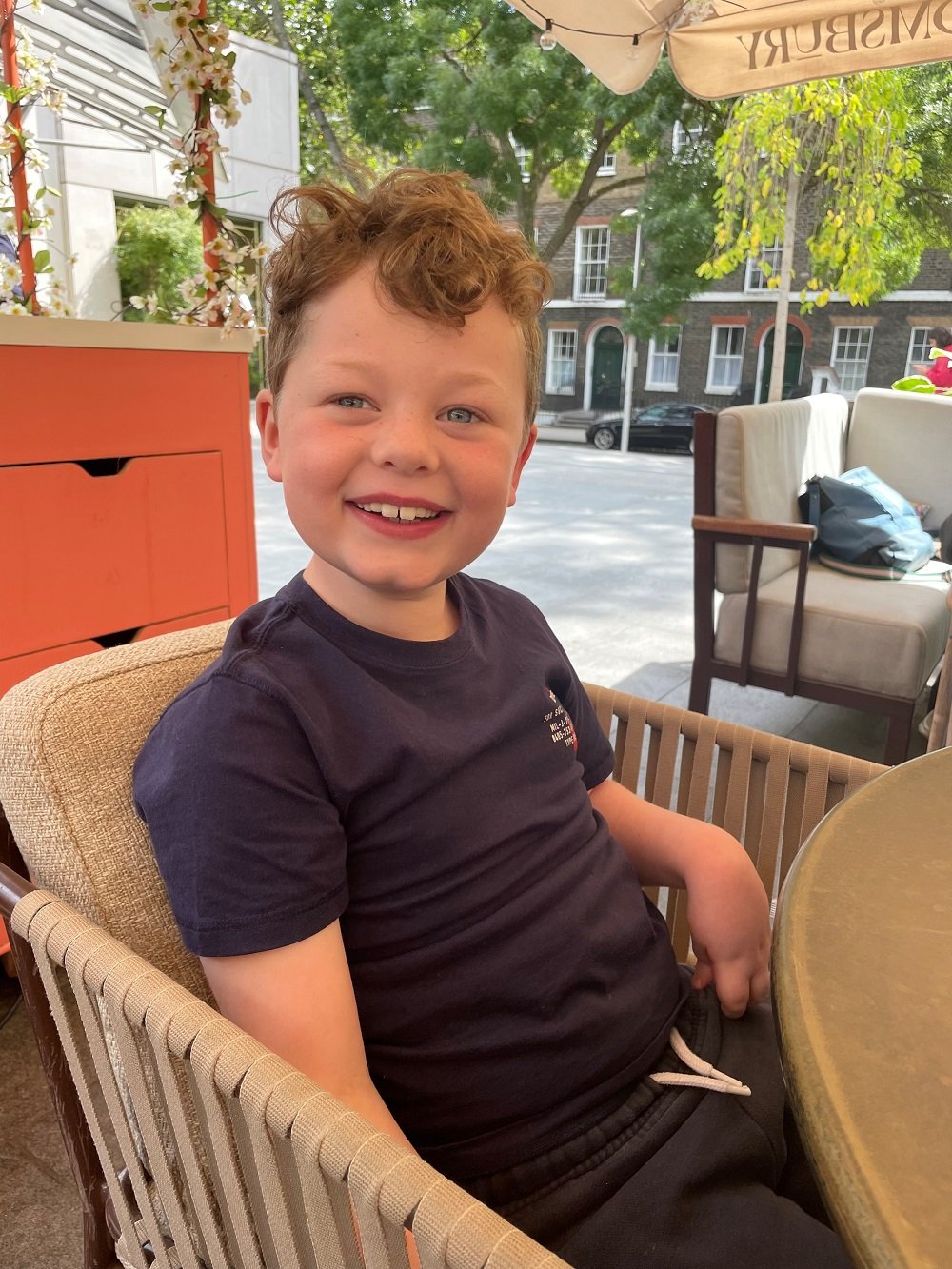 Sam is sitting in a wicker chair at a restaurant. He is smiling and looking at the camera.