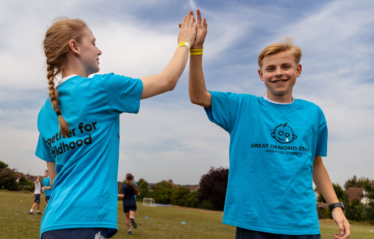 two teenagers, one boy and one girl, are high fiving. they are both in blue tshirts with the GOSH logo on the front. they are both smiling, with the boy looking into the camera. they are on a field
