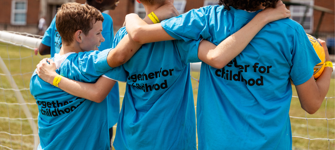 3 young boys with their arms around each other facing away from the camera, the boy on the left is smiling at the other two boys. they are all wearing blue thsirts with 'together for childhood' written on the back in black font. they are in front of a brick building