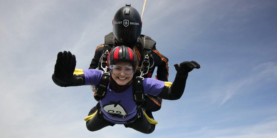 A woman wearing a GOSH Charity purple t-shirt, in the sky completing a skydive. It is a clear, sunny day, with blue skies.