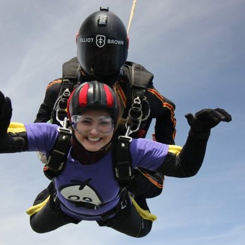 A woman wearing a GOSH Charity purple t-shirt, in the sky completing a skydive. It is a clear, sunny day, with blue skies.