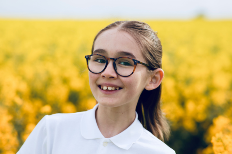 A teenager wearing glasses smiles to the camera. Behind her is a blurred, yellow floral background.