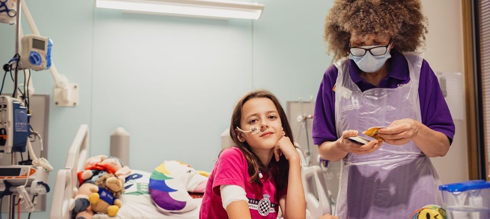 Photo of Sophia sitting down in her room at GOSH, looking to camera. Play specialist Lizzie stands next to her, holding and looking at play activities.