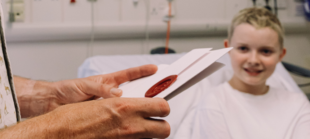 Hands holding GOSH Taskmaster envelope with wax seal. Girl in hospital room smiling in the background.