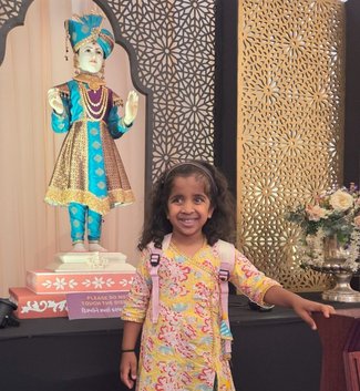 A small child in a white flowery dress smiles. She is standing in front of a statue at her temple.