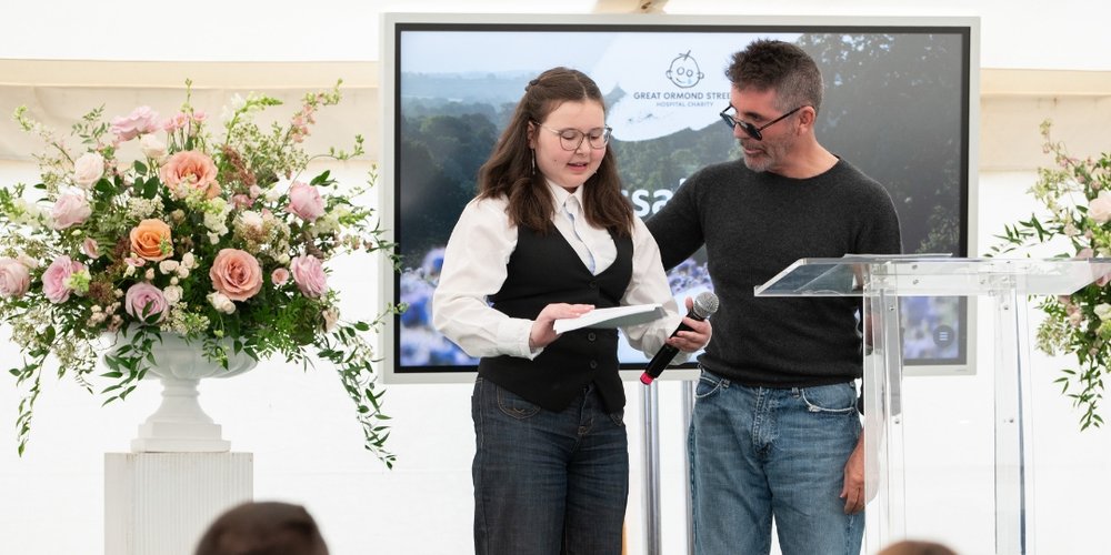 Simon Cowell pictured with GOSH patient Alyssa. They both stand on a stage. Alyssa has a microphone in her hand.