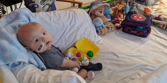 Photo of Max sitting on his bed at GOSH, surrounded by toys. Turning his head to look at his mum, who's taking the photo.