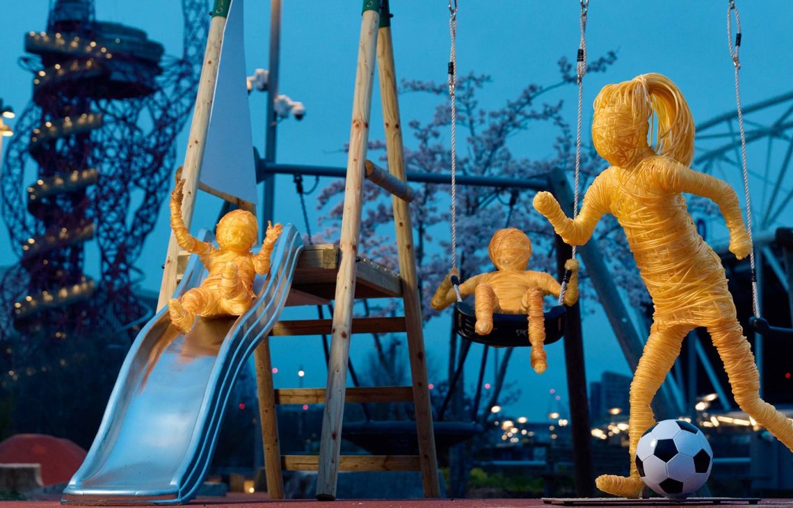 Three life-sized sculptures created using replica chemotherapy wires, situated in a park. One sculpture is going down a slide, another is on a swing and the other is kicking a football.